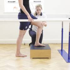 Side shot of a patient placing their left leg on a wooden box while holding on to two beams with a neurological therapist placing their left hand on the patients left knee.