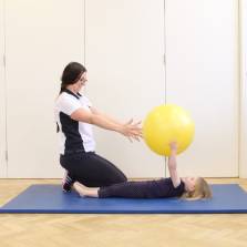 Side shot of a little girl lying on a blue mat holding a big yellow ball in the air and a paediatric therapist kneeling down with arms stretched out trying to grab the ball.