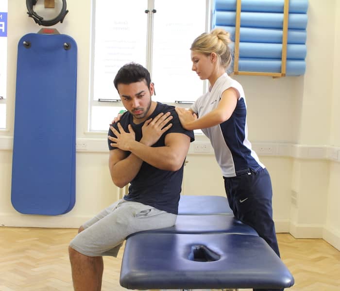 Side shot of man sat down with arms crossed over touching his shoulders at the front with therapist holding onto his back.