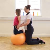 Side shot of little gitl knelt on a big orange ball with physiotherapist behind girl helping her.